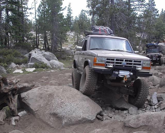 Lifted tan Ford Bronco II with front winch and roof rack crawling rocks on a pine trail