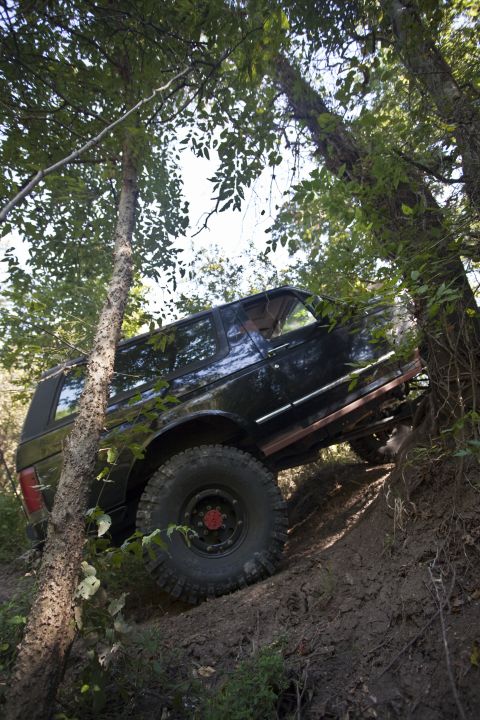 1995 Ford Bronco - A lifted black two-door SUV tips at a steep angle on a wooded dirt embankment, shot from below through saplings. It rides on tall mud-terra…