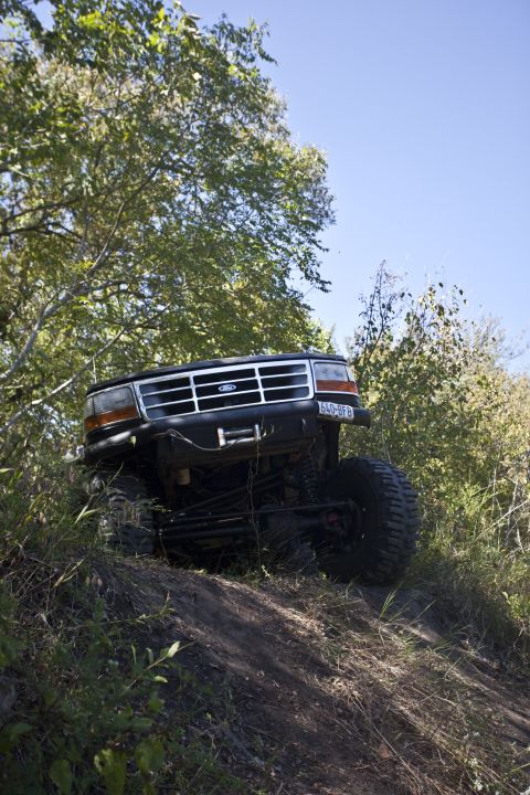 1995 Ford Bronco - A lifted black Ford Bronco navigates an off-road trail with dense foliage surrounding it.