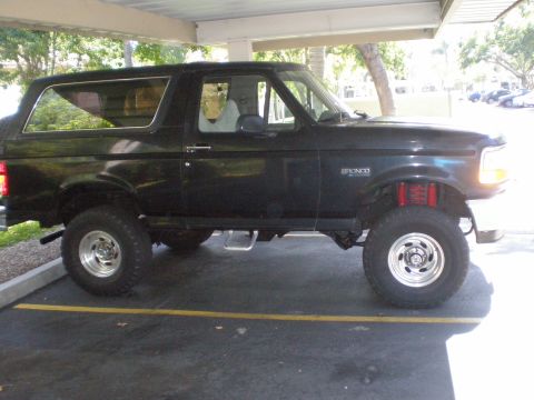 1995 Ford Bronco - This black Ford Bronco is lifted with red shocks and parked under a covered area.