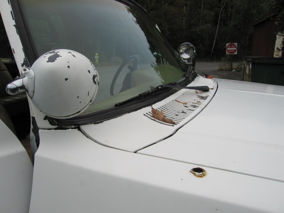 White truck hood with peeling paint on the side mirror and rust around a hole