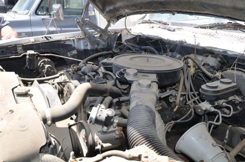 1987 Dodge 1500 - The engine bay of a weathered truck, with visible engine components and a second vehicle in the background.