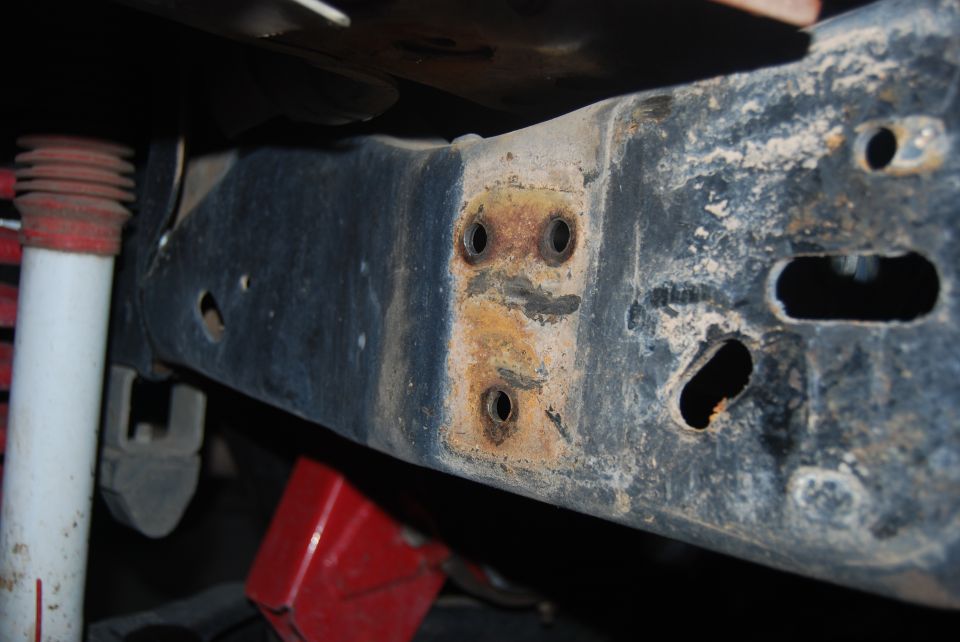 Close-up of a rusted vehicle frame rail with mounting holes and a white shock absorber visible