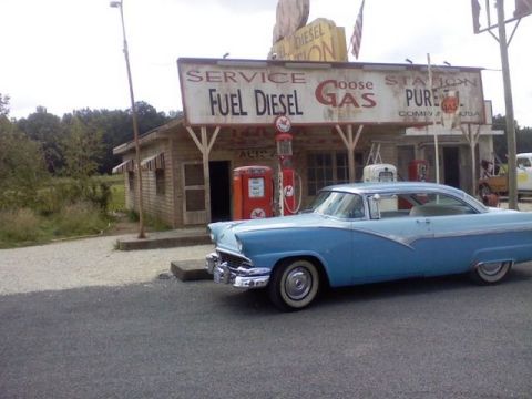 1991 Ford LTD Crown Victoria - A classic blue 1956 Ford Fairlane coupe is parked in front of a vintage gas station, complete with old-fashioned pumps and signage.