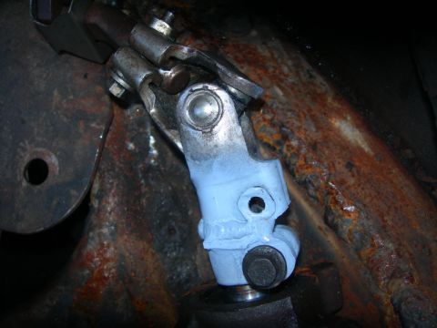 1984 Ford Bronco - A close-up view of a steering linkage component, painted light blue, showing signs of wear and rust on surrounding metal parts.