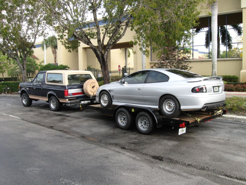 Two-tone Ford Bronco towing a silver coupe on an open car trailer in a parking lot