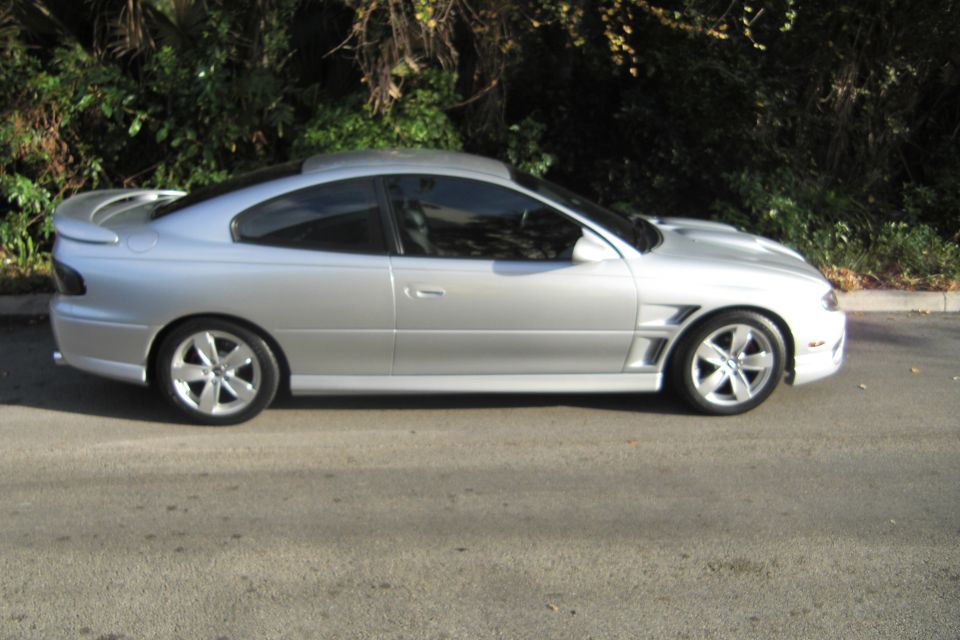 Side view of a silver Pontiac GTO coupe with rear spoiler and alloy wheels parked by greenery