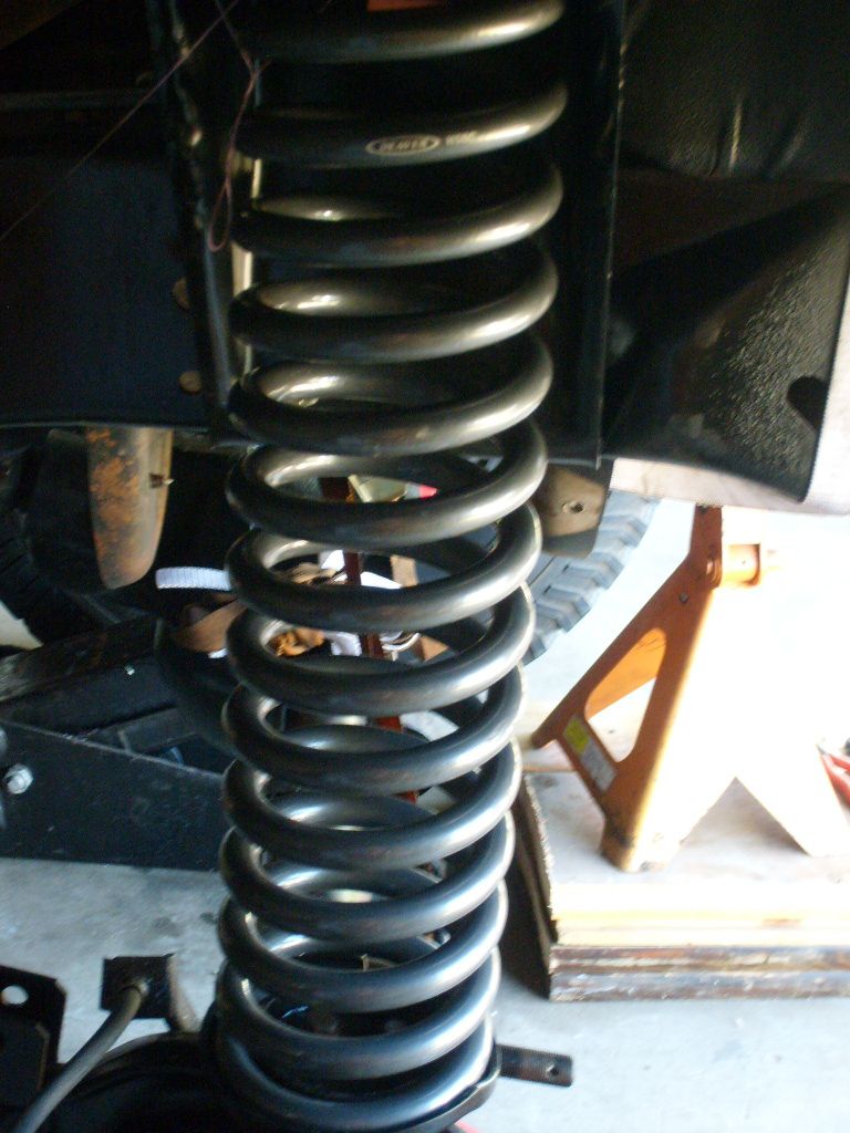 close-up of a large coil spring on a vehicle's suspension, with a jack stand visible in the background