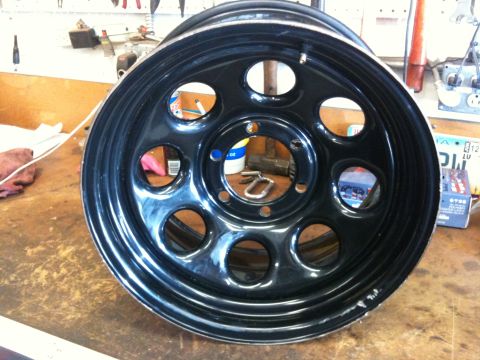 2009 Suzuki Samurai - A close-up of a black steel wheel, likely for an off-road vehicle, resting on a wooden surface.