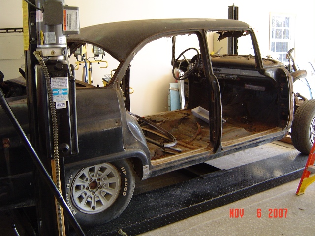 Black 1955 Ford body on a lift, interior removed, rust visible, white aftermarket wheels