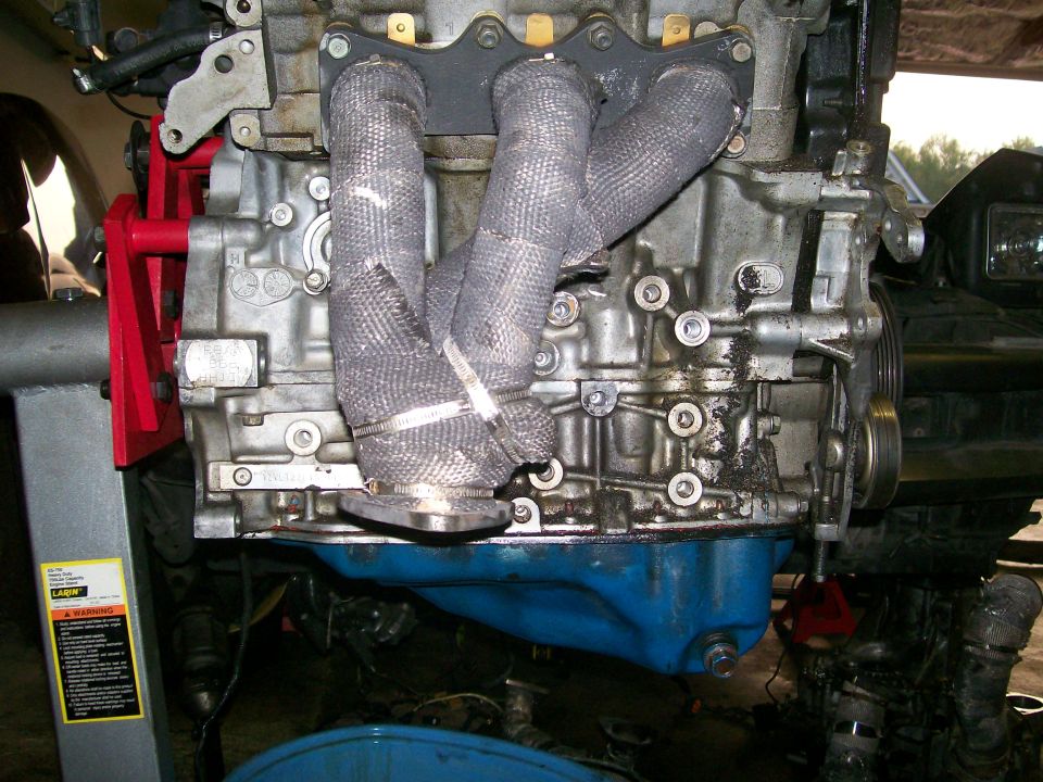 Engine block with heat-wrapped exhaust manifold, supported by a red engine hoist, with a blue oil pan