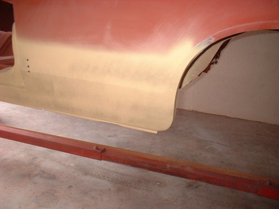 Car body panel being prepped for paint, showing primer and red paint. A red metal support beam is in the foreground