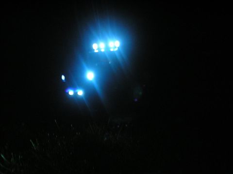 1994 Ford Escort - A vehicle is illuminated by bright blue auxiliary lights in the dark, with grass visible below.