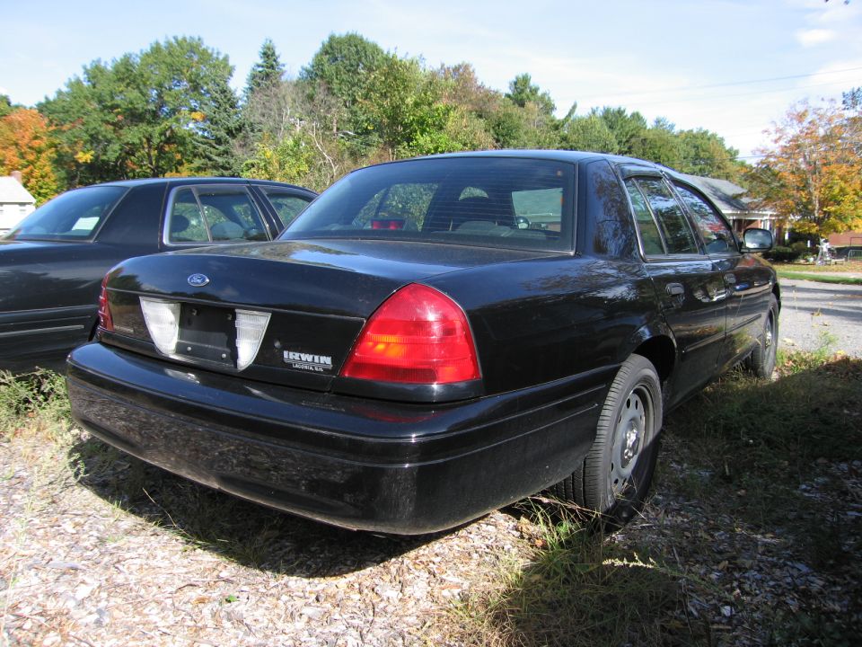 Black Ford Crown Victoria, rear 3/4 view. Another black car is partially visible to the left. The car has a dented rear bumper