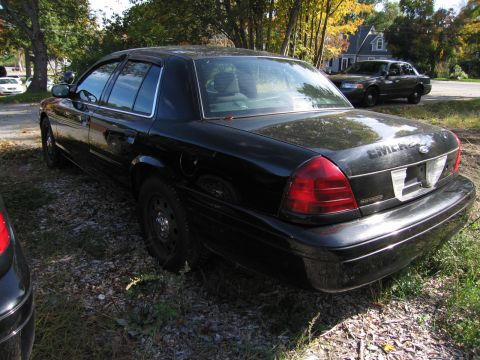 1991 Ford LTD Crown Victoria - Two black sedans are parked on a gravel lot with trees and a house in the background.