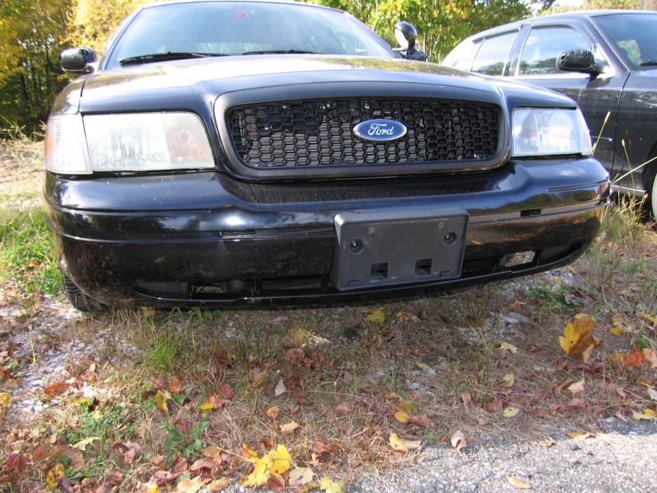 Black Ford Crown Victoria front grille and headlights, parked outdoors with autumn leaves on the ground