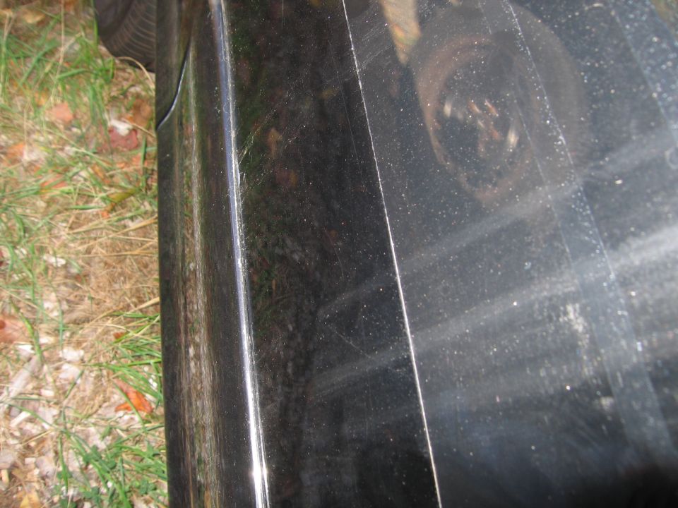 Close-up of a black car's side panel and wheel well. Grass and dirt are visible on the left