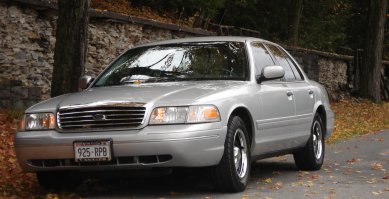 2000 Ford Crown Victoria - A silver 2000 Ford Crown Victoria is parked on a road with fall foliage and a stone wall in the background.