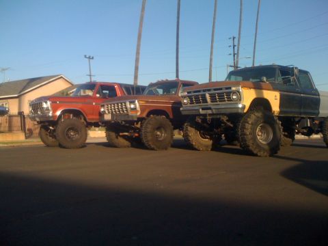 1979 Ford Bronco - Three lifted pickup trucks are parked in a row on a sunny day. The front truck is orange, the middle is brown, and the rear is black and ye…