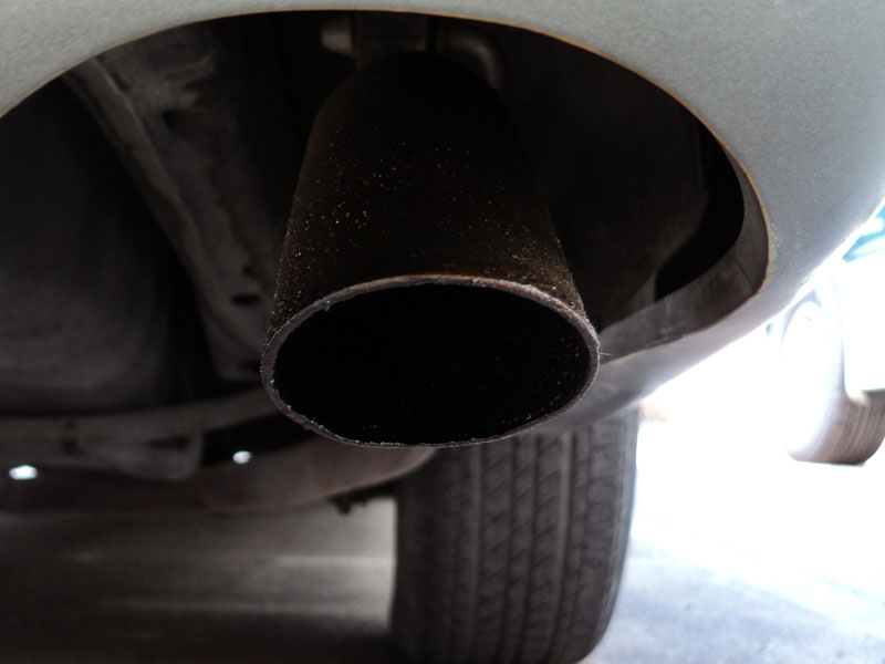Angle-cut sooty exhaust tip exiting under a silver car's rear bumper behind the tire