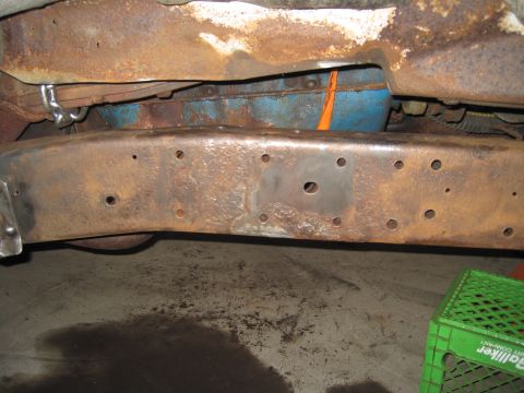 1979 Ford Bronco - Underbody view of a rusty steel frame crossmember with multiple drilled holes above a concrete floor.