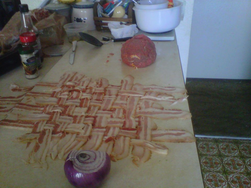 Woven bacon strips and a red onion on a countertop with kitchen items in the background