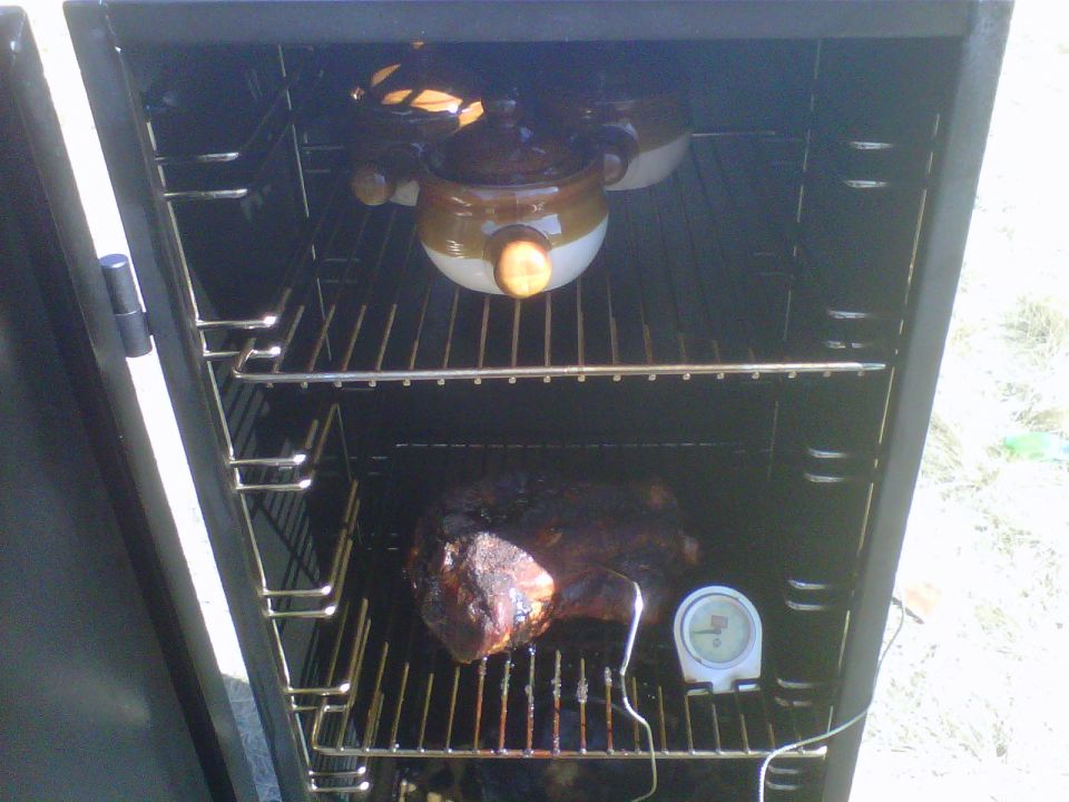 Inside of a smoker with a large piece of meat on a rack and two pots on an upper rack. A thermometer is next to the meat