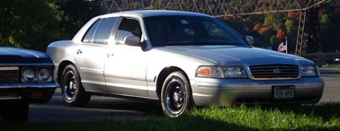 2000 Ford Crown Victoria - A silver 2000 Ford Crown Victoria is parked on the grass, with another vehicle partially visible to its left.