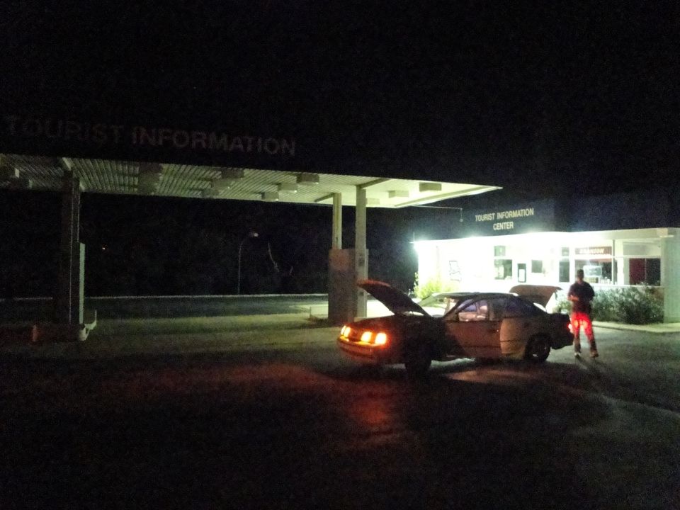 Yellow sedan with hood and trunk open at night, under a covered structure with "TOURIST INFORMATION" text