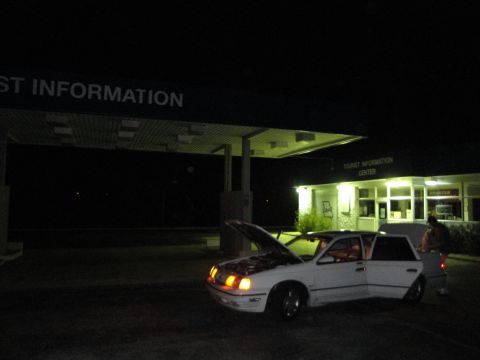 1989 Ford Taurus - A white 1989 Ford Taurus sits with its hood and trunk open at a tourist information center at night.
