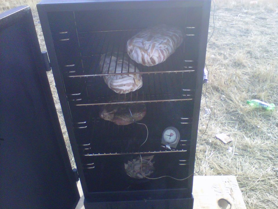 Open smoker cabinet with racks holding food wrapped in bacon and other items; a thermometer is visible