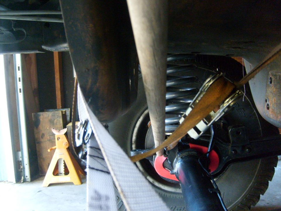 Underneath a vehicle, showing a coil spring, exhaust pipe, and a strap holding something. A yellow jack stand is visible