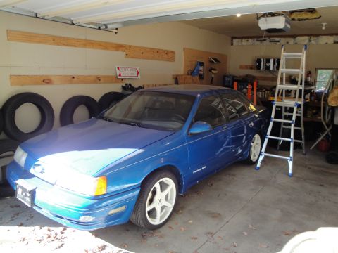 1989 Ford Taurus - A blue 1989 Ford Taurus sedan is parked inside a garage, with a ladder and tires visible nearby.