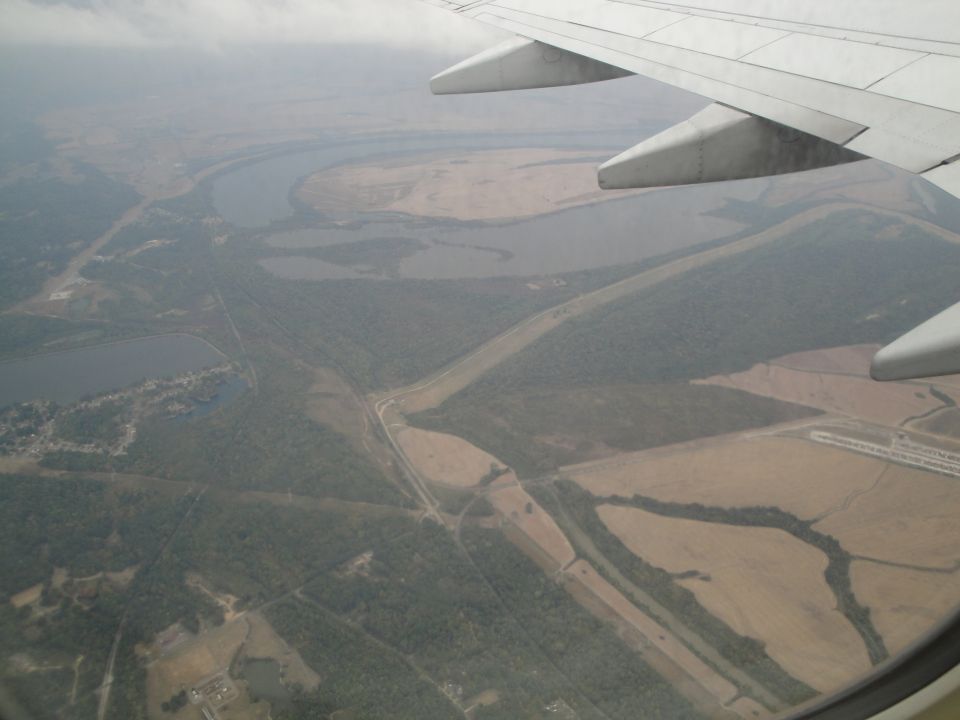 View from airplane window shows a winding river, fields, and trees below, with the wing of the plane in the foreground