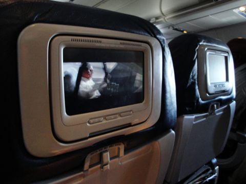1989 Ford Taurus - A view inside an airplane cabin shows two seatback entertainment screens, each displaying a different movie.