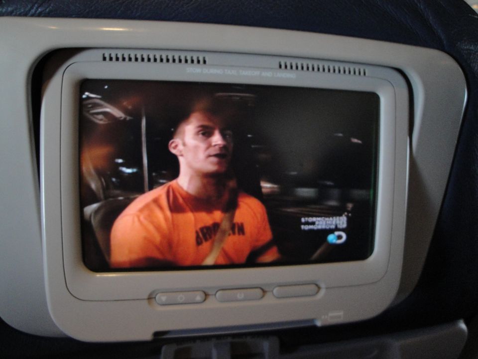 Man in orange shirt with 'BROOKLYN' visible, driving a car at night. Discovery Channel logo in corner