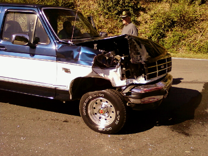 Front end collision damage on a blue and white Ford F-Series pickup truck. Grille and hood are heavily damaged