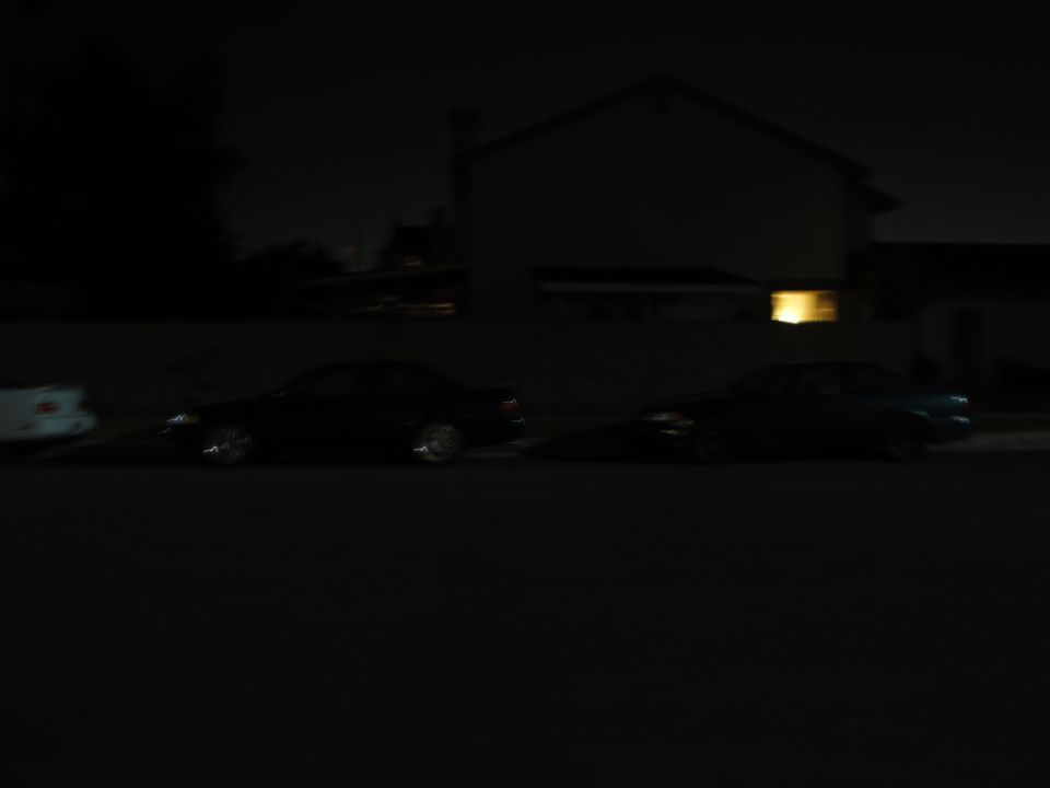 Dark sedan parked on street at night, side view. Another car is partially visible to its right