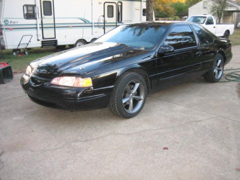 1996 Ford Thunderbird - A black 1996 Ford Thunderbird coupe is parked on a concrete driveway next to a white RV and a white pickup truck.