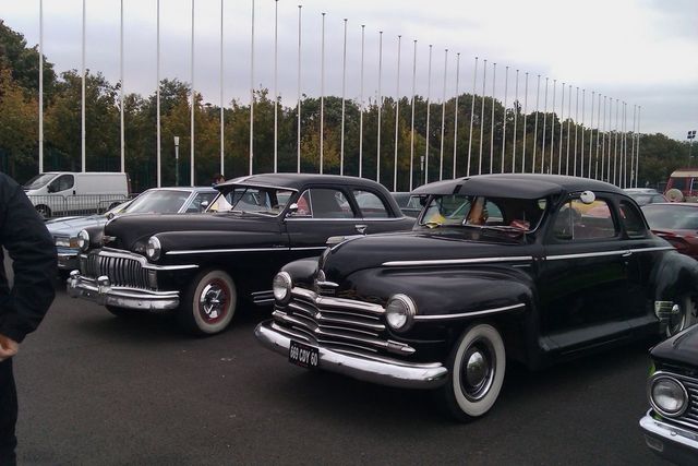 Two black classic coupes parked outdoors, one in front of the other. A white van is visible in the background
