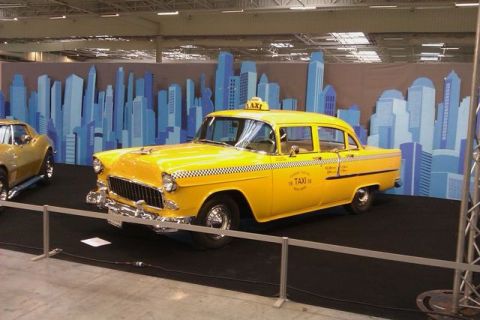 1991 Ford LTD Crown Victoria - A yellow 1955 taxi cab is displayed in front of a New York City skyline backdrop, with another car visible to the left.