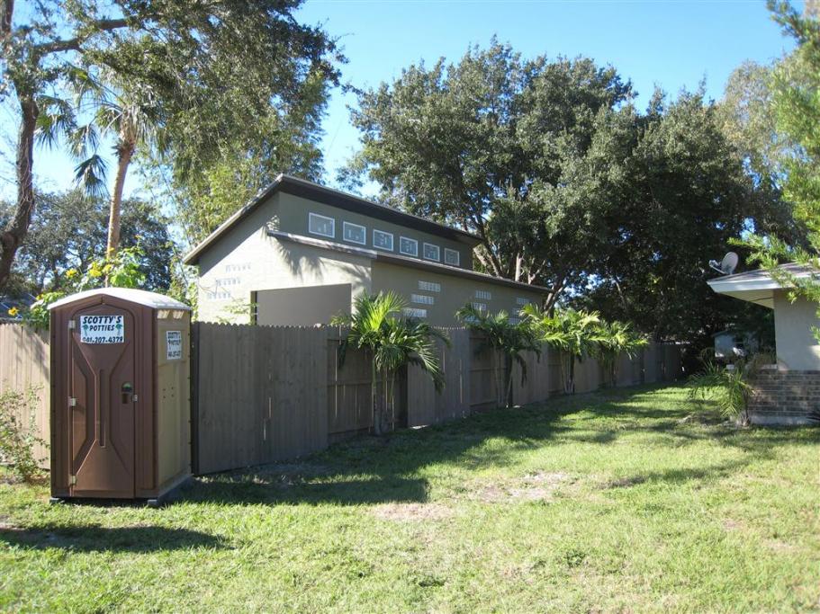 Brown portable toilet next to a wooden fence and a building