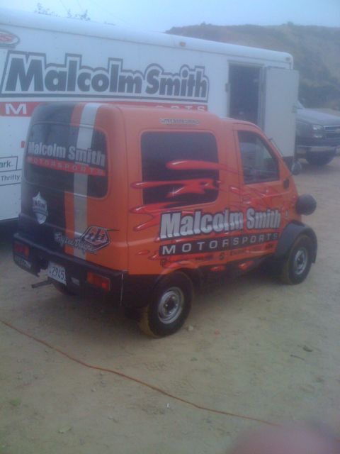 1968 Ford Mustang - An orange van with racing decals is parked in front of a large white trailer with "Malcolm Smith" branding.