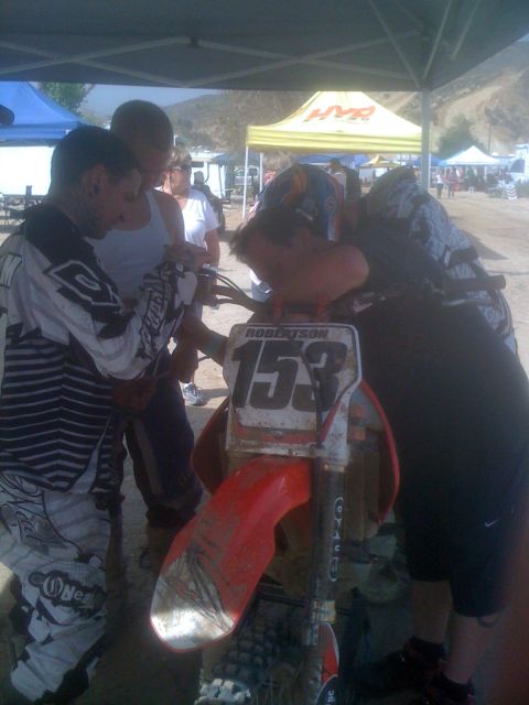 1968 Ford Mustang - Riders inspect a dirt bike at an outdoor event. The bike has a number plate and red fender.