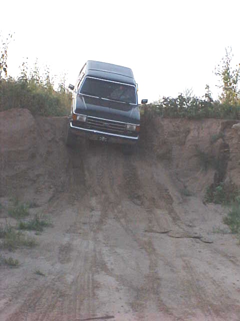 1989 Ford Bronco - A dark SUV navigates a steep dirt incline, showing its front grille and tires on the loose terrain.