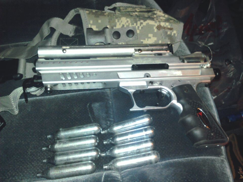 Silver paintball marker with CO2 cartridges and camo harness on a gray cloth seat
