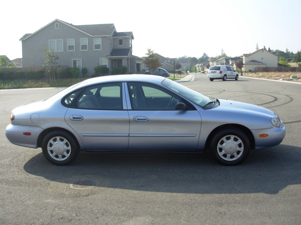 Silver sedan parked on a residential street, side view. Shows a white minivan in the background