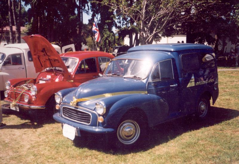 Blue Morris Minor panel van with gold signwriting at a grass car show, red Minor behind