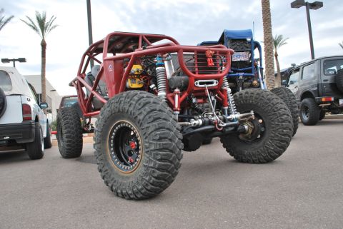 Suzuki Samurai - Low front three-quarter view of a red tube-chassis rock buggy parked on asphalt among other 4x4s, showing its full exo cage, exposed radiat…