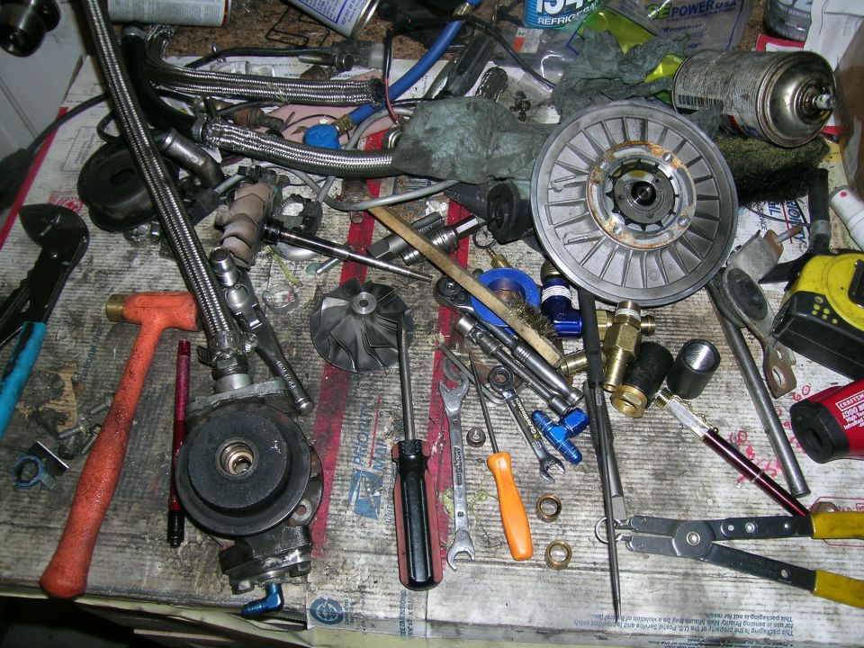 Pile of car parts and tools on a workbench, including a turbocharger, hoses, and wrenches
